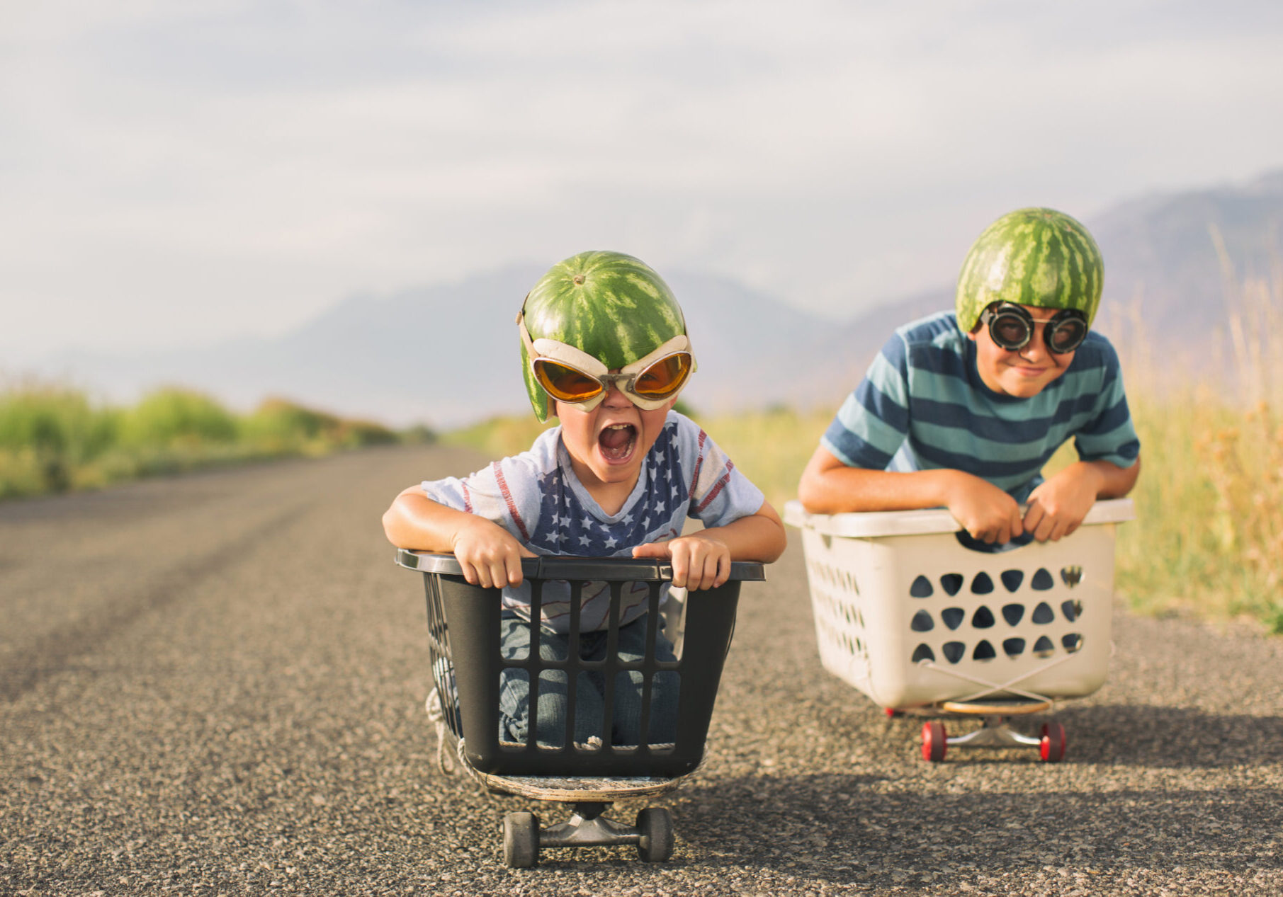 A young boy races his brother in a makeshift go-cart while wearing watermelon helmets and goggles. He is excited as he is winning the race.